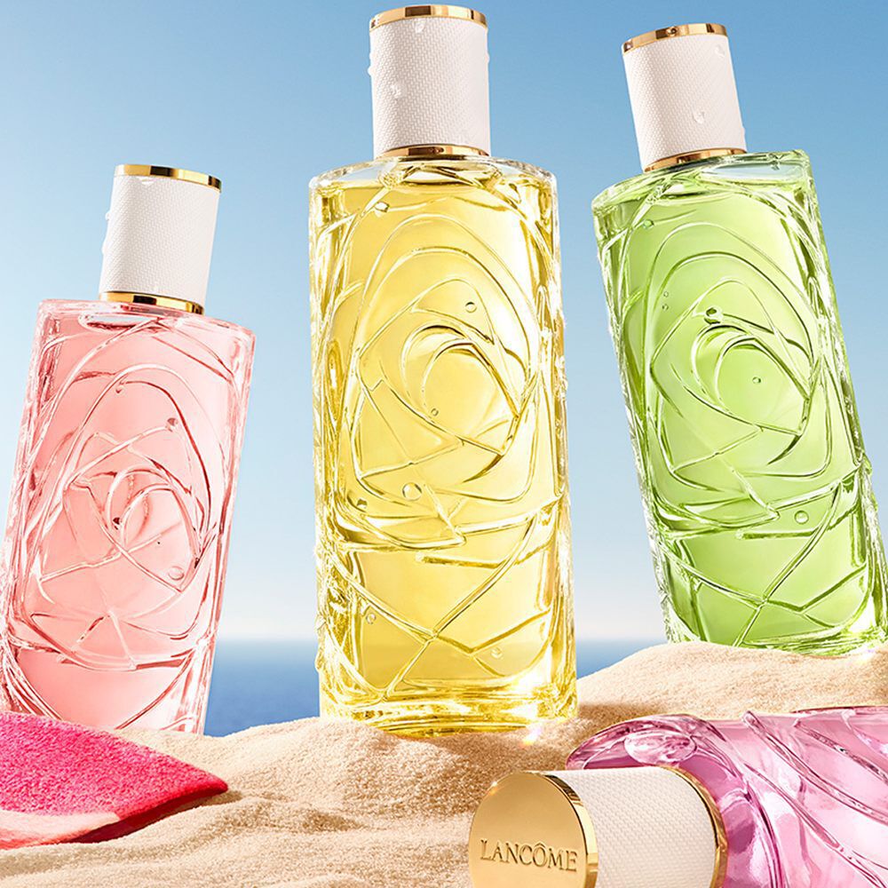 Beach scene with three upright perfume bottles (pink, yellow, green) and one pink bottle lying in the sand. A pink-striped towel is on the left with a clear blue sky and ocean horizon in the background.