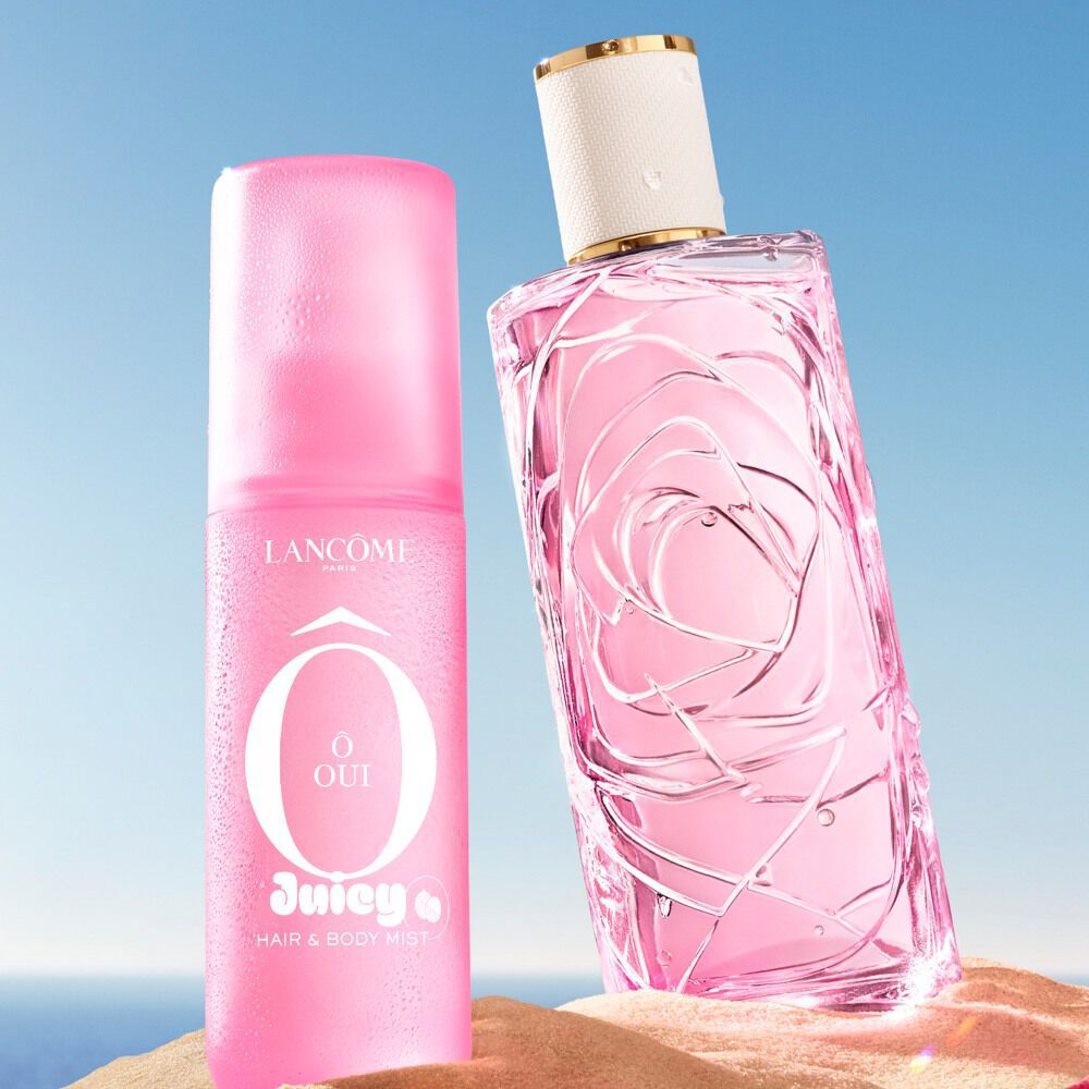 Two pink bottles, one textured glass perfume and one spray, standing on a sandy beach next to a folded pink-striped towel. Clear blue sky and ocean in the background