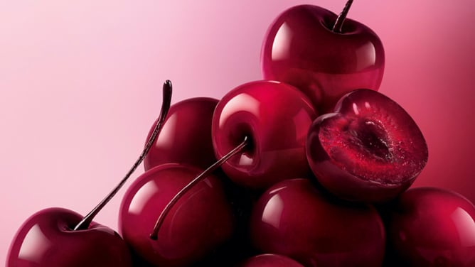 A pile of glossy, dark red to black cherries fills the bottom and right of the frame, set against a soft pink background.